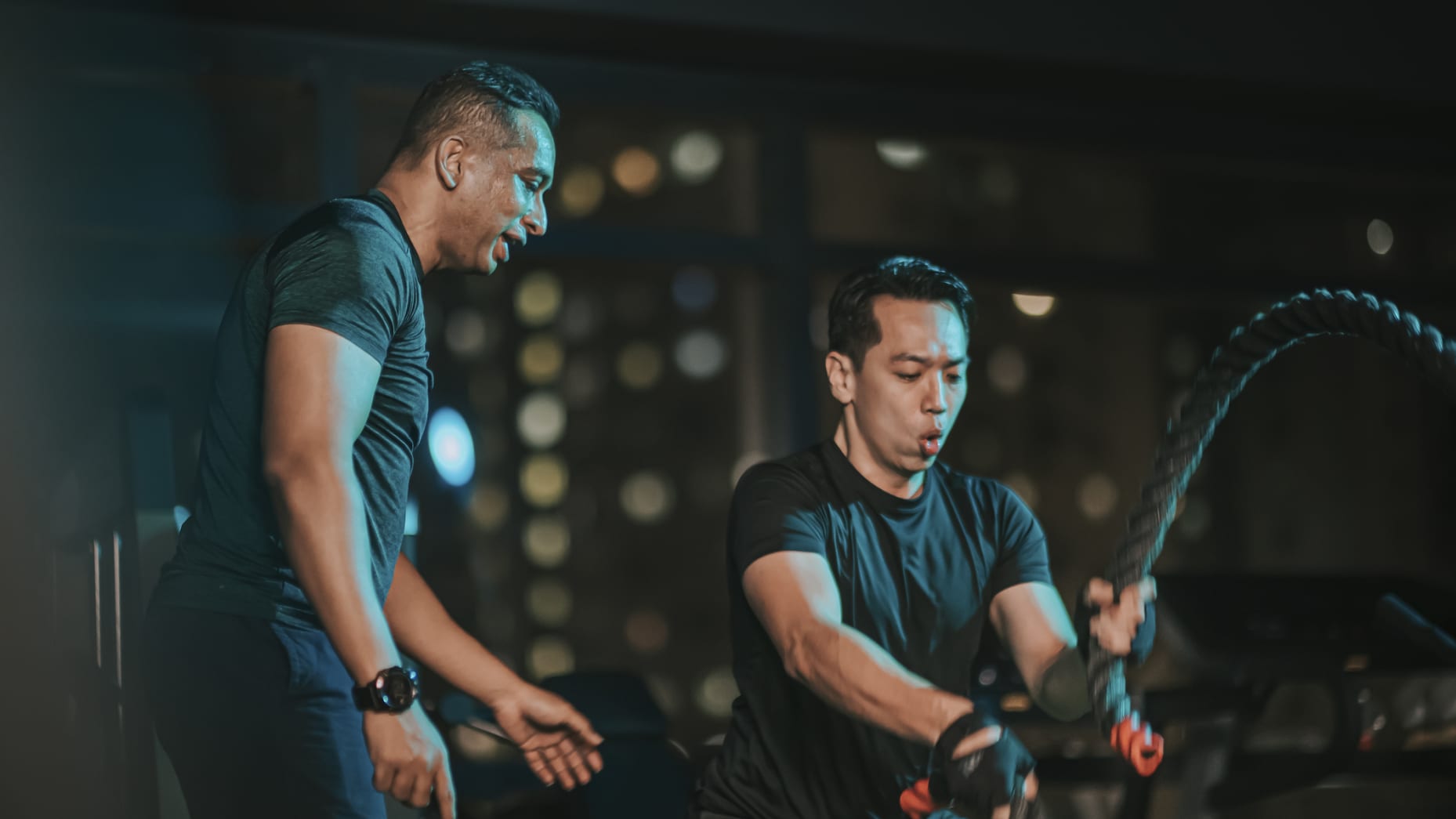 Body Positive Asian Chinese Mid Adult woman practicing battle rope in gym with her indian male personal trainer