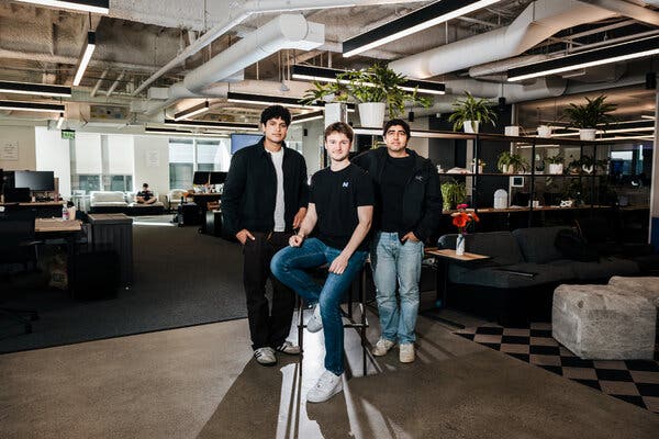 Mr. Hiremath and Mr. Midha stand on either side of Mr. Foody, who is sitting on a stool in a spacious room under a ceiling lined with tubing.