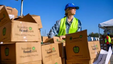 Getty Images A woman wearing a blue baseball cap and a hi-viz vest stands outdoors and looks to the right. In fron of her is a pile of boxes that read 'Houston Food Bank'