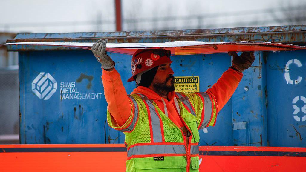 A bearded construction worker wearing an orange sweatshirt and yellow safety vest carries a sheet of construction material over his his head. In the background there is a a blue container. The picture was taken at the site of the Gateway Tunnel construction project as seen from North Bergen, New Jersey, on February 10, 2026.