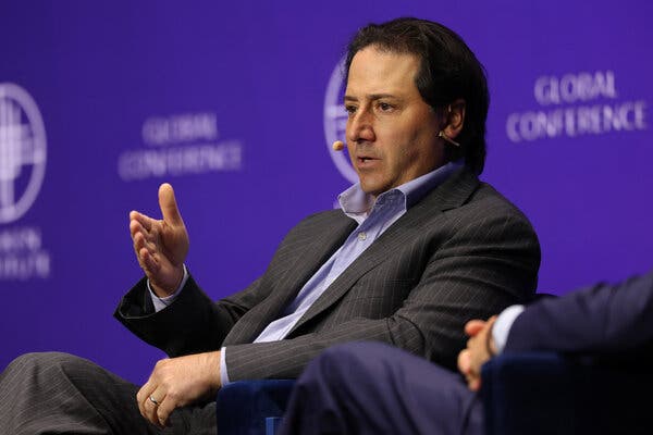 Michael Arougheti, wearing a headset microphone and sitting on a stage, gestures while speaking