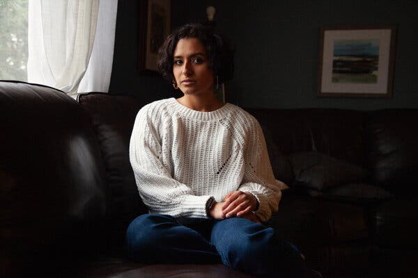 A portrait of Erin Torres, wearing a white sweater and jeans, sitting on a couch in a darkened room near a window.